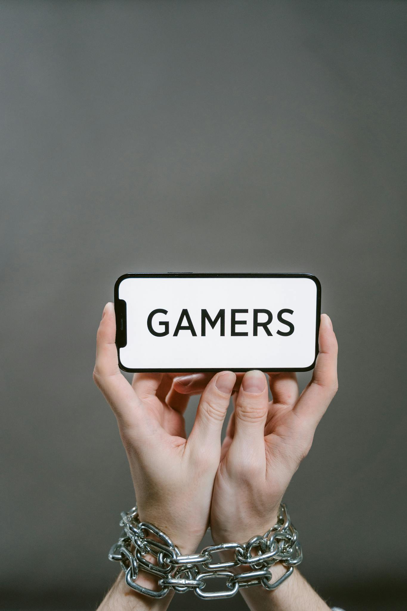Person's hands in chains holding a smartphone displaying the word 'GAMERS' on a gray background.